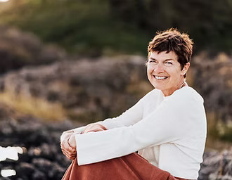 A woman with short hair wearing a white sweater and orange skirt smiles into the camera. She is is sitting outside on rocks, seen out-of-focus in the background.