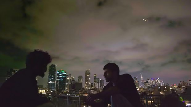 Two men talking on a rooftop in the dark. The sky behind is lit by the lights from the city in the background.