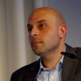 A bald man wearing a suit jacket. He is staring past the camera to the left.