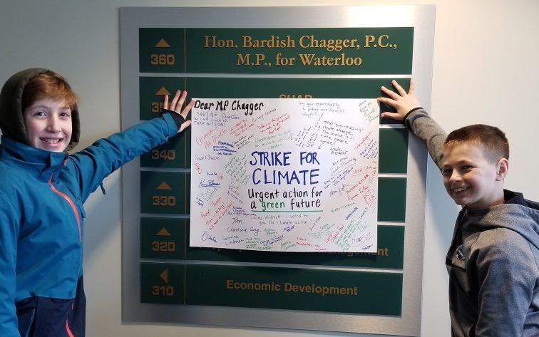 Two students holding a poster with signature over the office sign for Hon. Bardish Chagger, P.C., M.P., for Waterloo